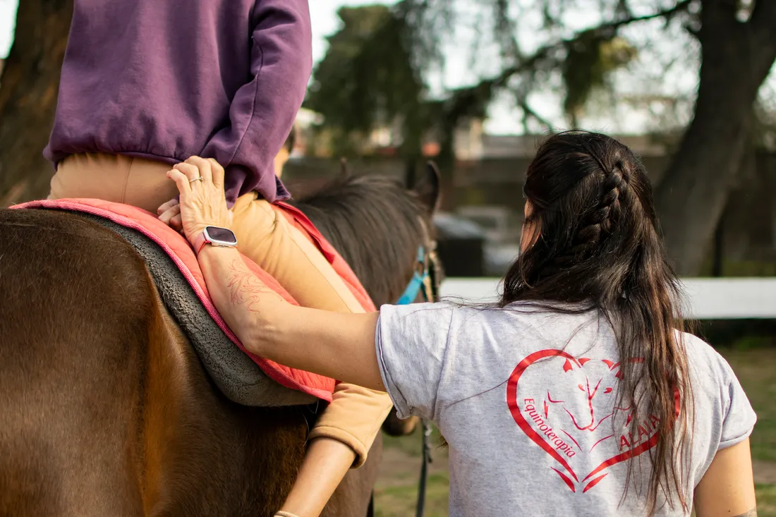 Una profesional acompaña con su mano a una paciente que monta a caballo