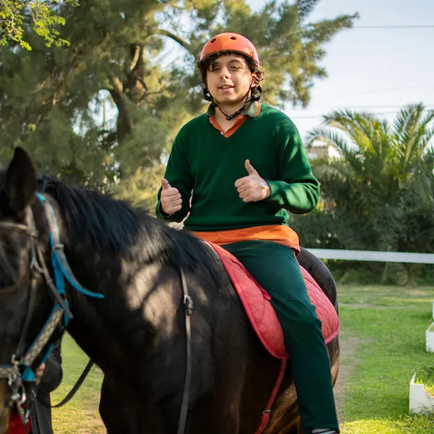 Un paciente con casco sonríe y levanta los pulgares, arriba de uno de los caballos